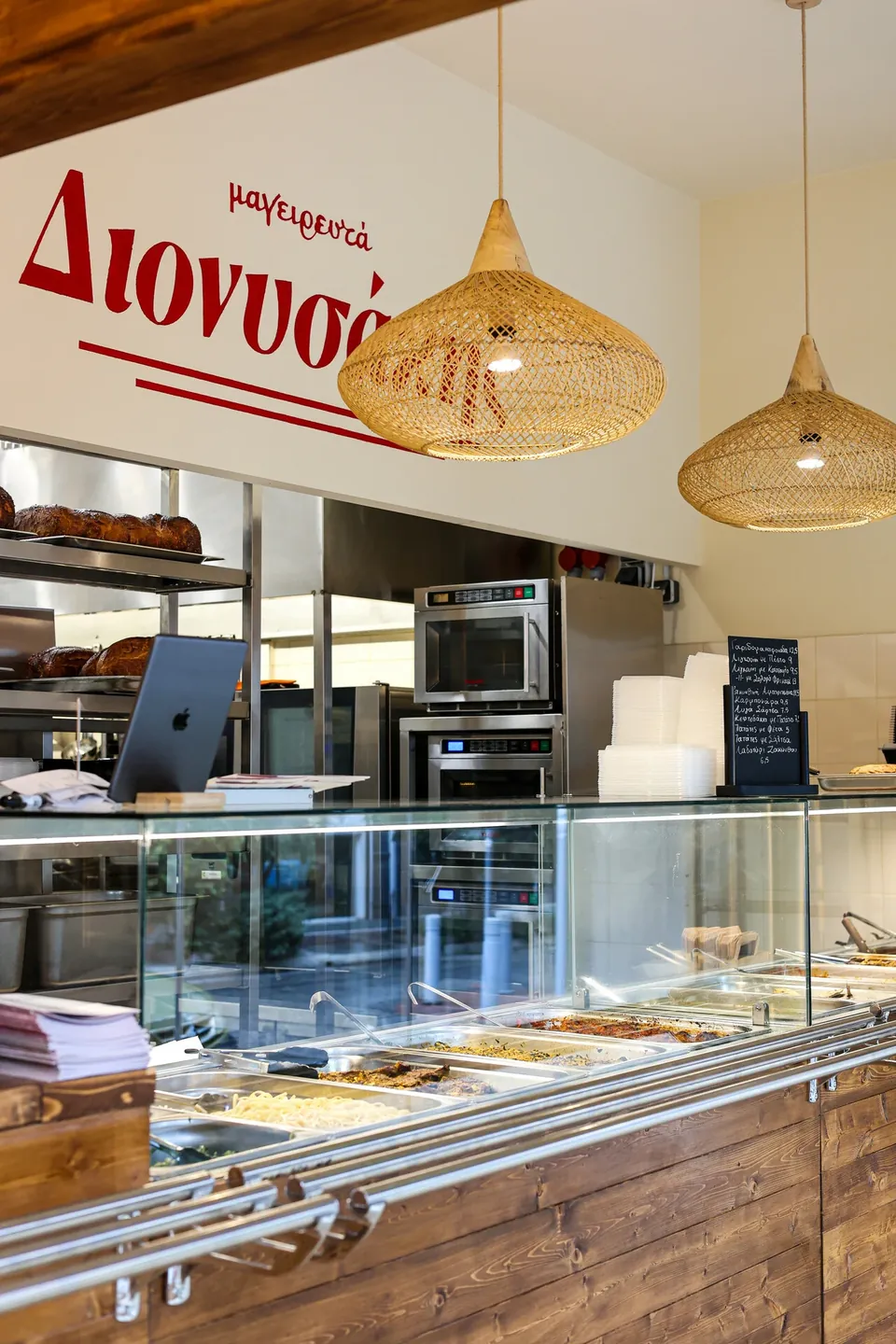 Η βιτρίνα του Διονυσάκη με τα ταψιά της ημέρας και πίσω η ανοιχτή κουζίνα με το logo στον τοίχο